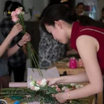 Asian woman wear sleeveless maroon blouse arranging fresh flowers on a table, flower arranging workshop, flower arrangement class​